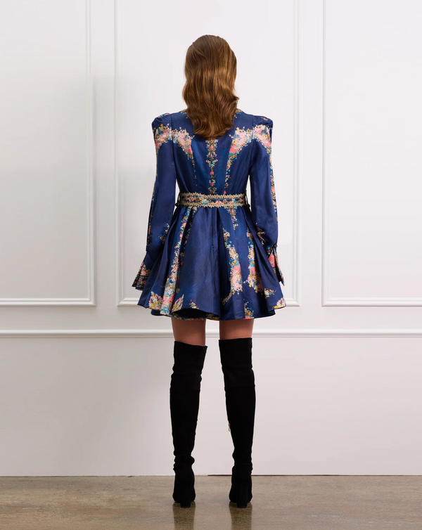 Woman wearing a blue floral dress with long sleeves and black knee-high boots against a white paneled wall.