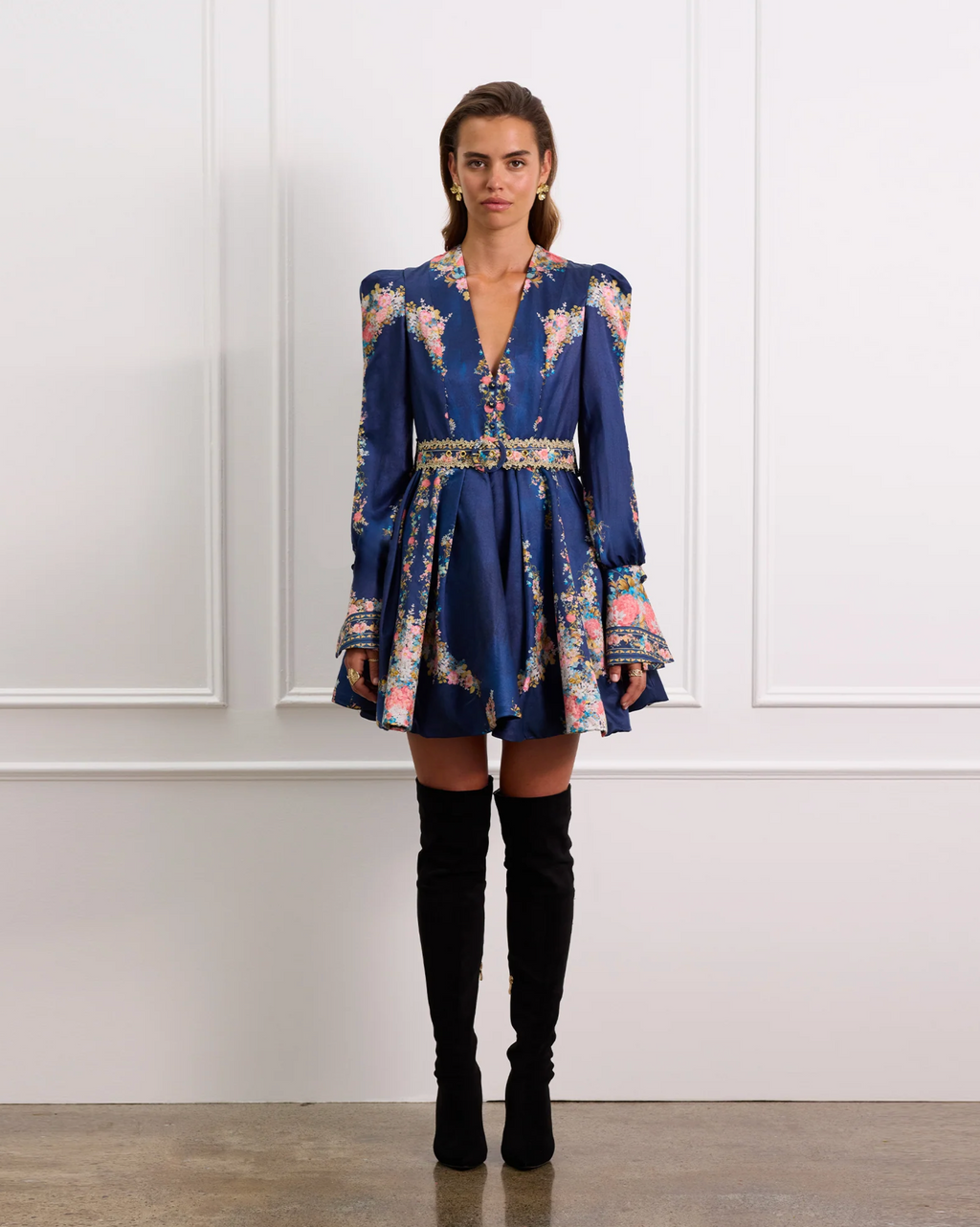 Woman wearing a blue floral dress with black knee-high boots against a white paneled wall.